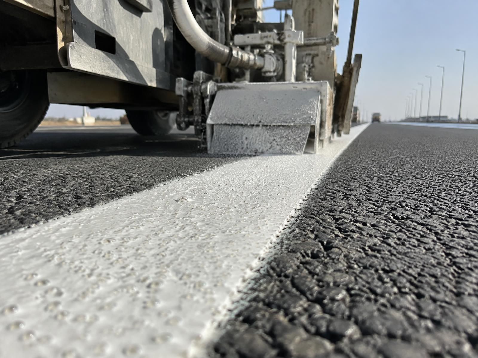 A close view along a freshly laid white road line, the marking machine’s shoe still above it and glass beads scattered across the wet paint, coarse black asphalt to one side and a line of lamp posts running to the horizon.